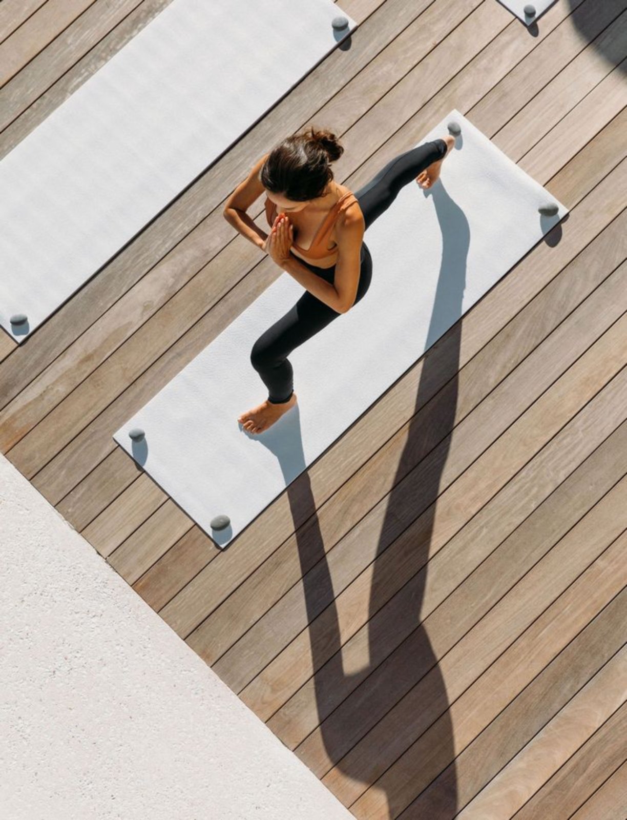 A woman balancing on one leg on a yoga mat on a sunlit wooden deck, seen from above